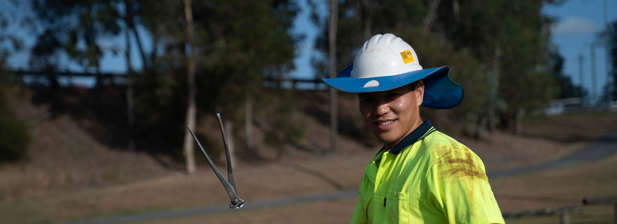 In a vibrant yellow work uniform and a unique hybrid hard hat/baseball cap, this cheerful individual, possibly engaged in labour hire services, is outdoors wielding tongs. Behind them lies a picturesque scene of trees and lush grass beneath the clear blue sky.