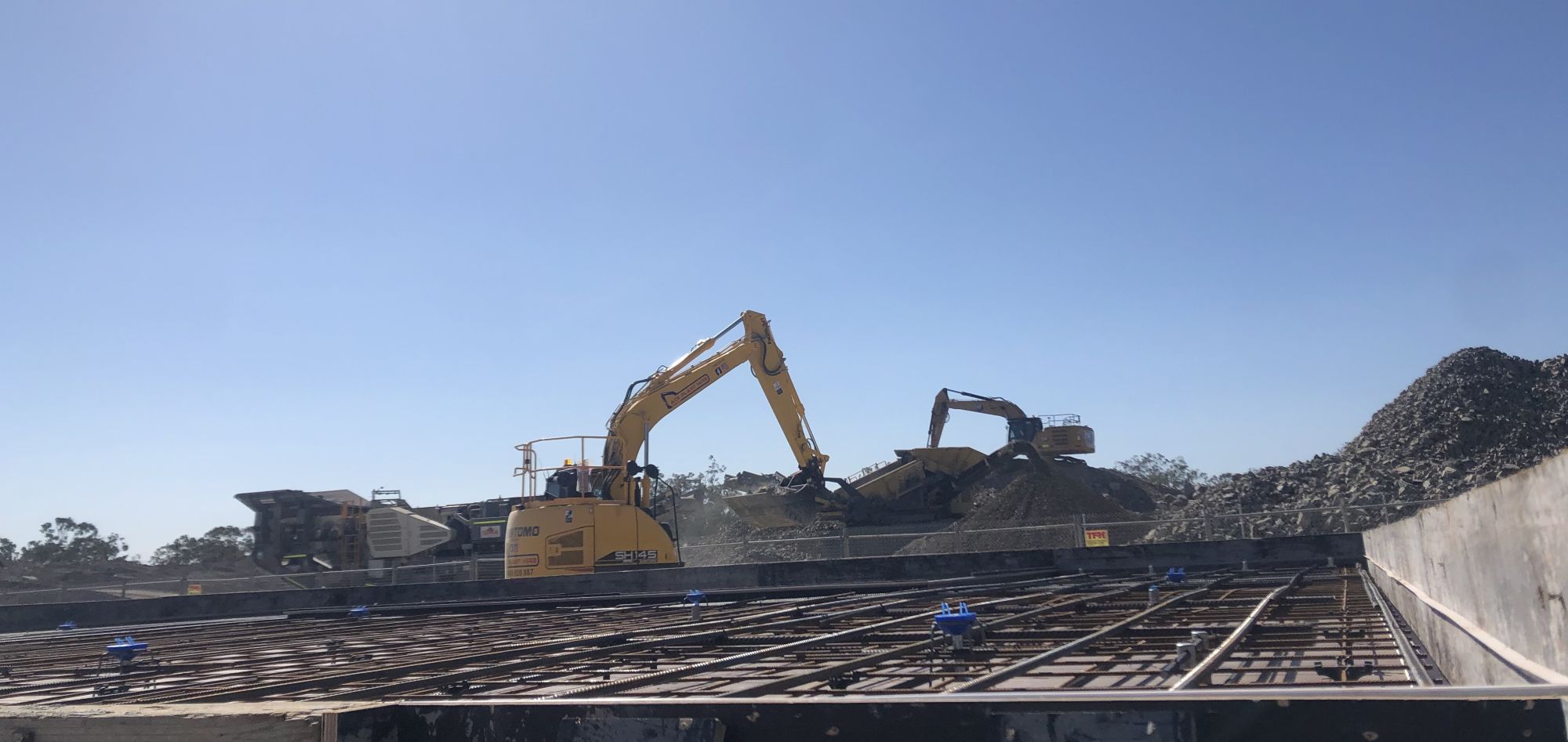 A construction site with two large yellow excavators working on piles of gravel showcases seamless efficiency, thanks to reliable labour hire services. A metal rebar structure stands in the foreground under the clear blue sky, highlighting a bright, sunny day.