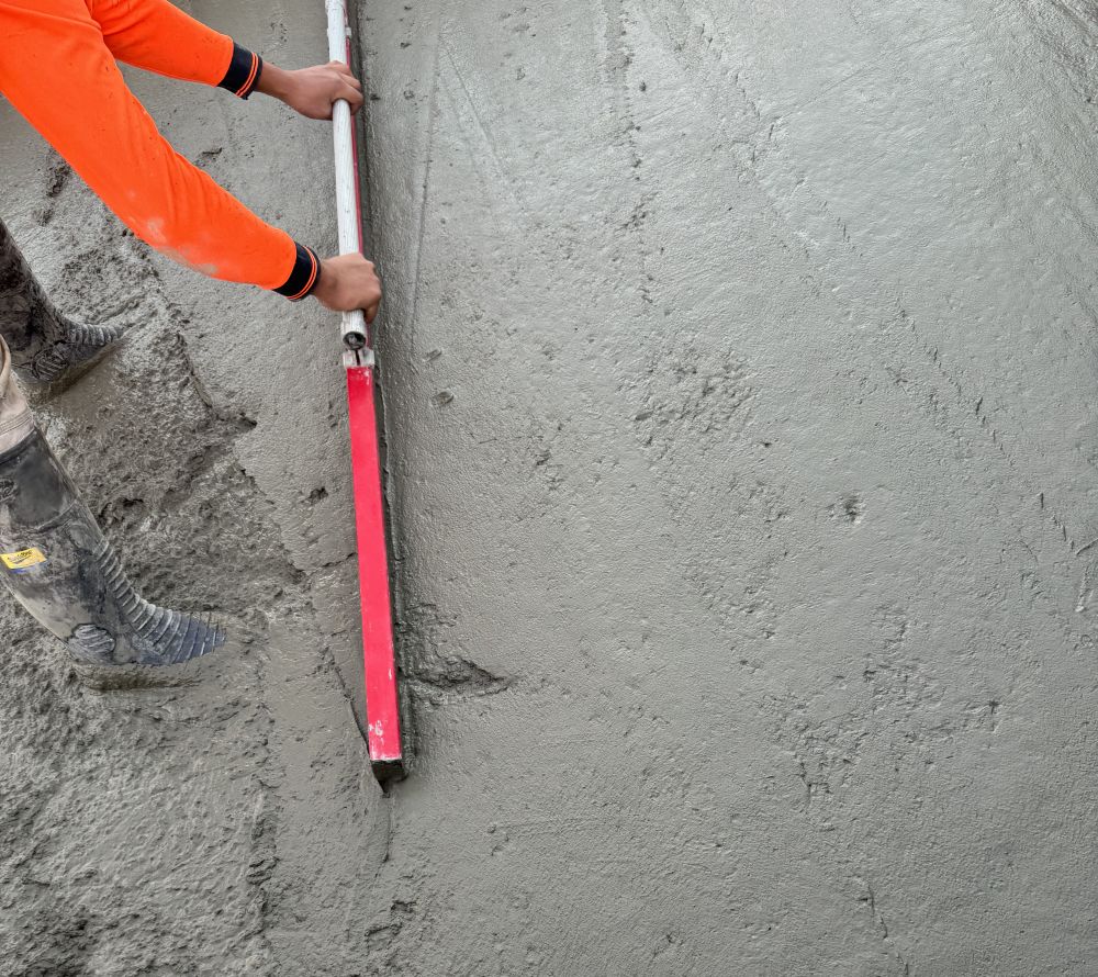 A person wearing an orange shirt and rubber boots, possibly from a labour hire service, is using a long, red-handled tool to smooth a freshly laid concrete surface. The concrete appears wet and even.