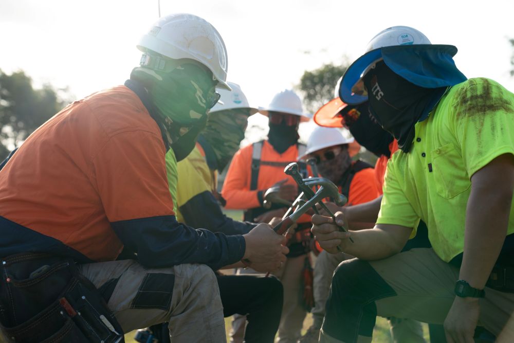 A group of construction workers in high-visibility safety gear and helmets gather outdoors, discussing plans. With sunlight filtering in the background, they appear focused and ready for action, embodying the spirit of efficient labour hire services.