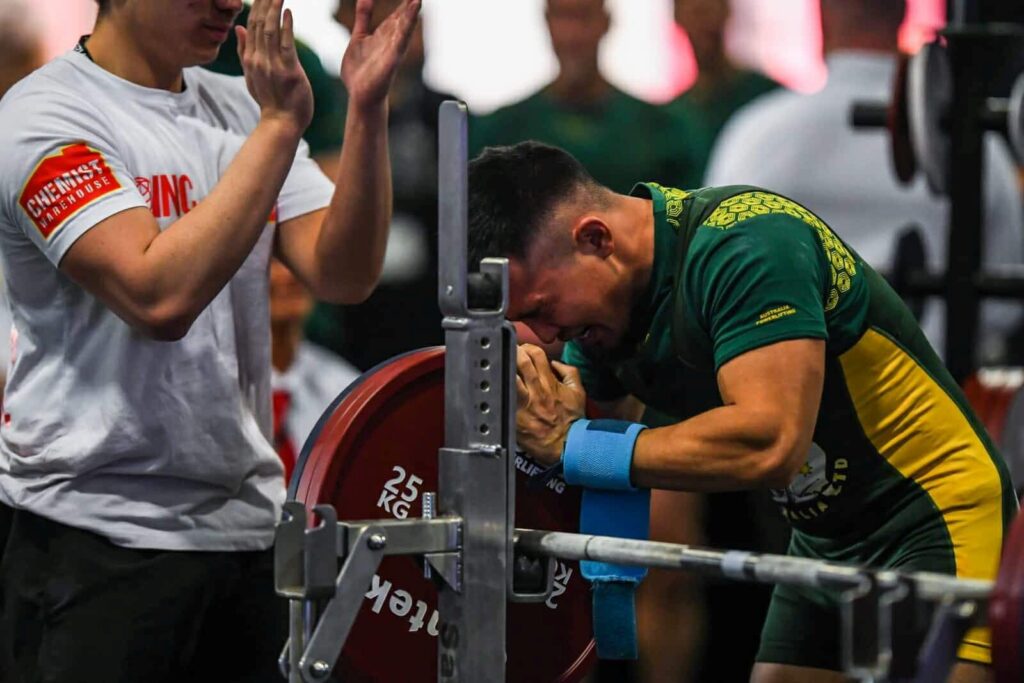 A weightlifter in a green and yellow outfit grips a bar loaded with heavy weights, appearing strained but focused. A supporter in a white shirt claps nearby, much like labour hire services providing crucial support to help the athlete achieve their lift.