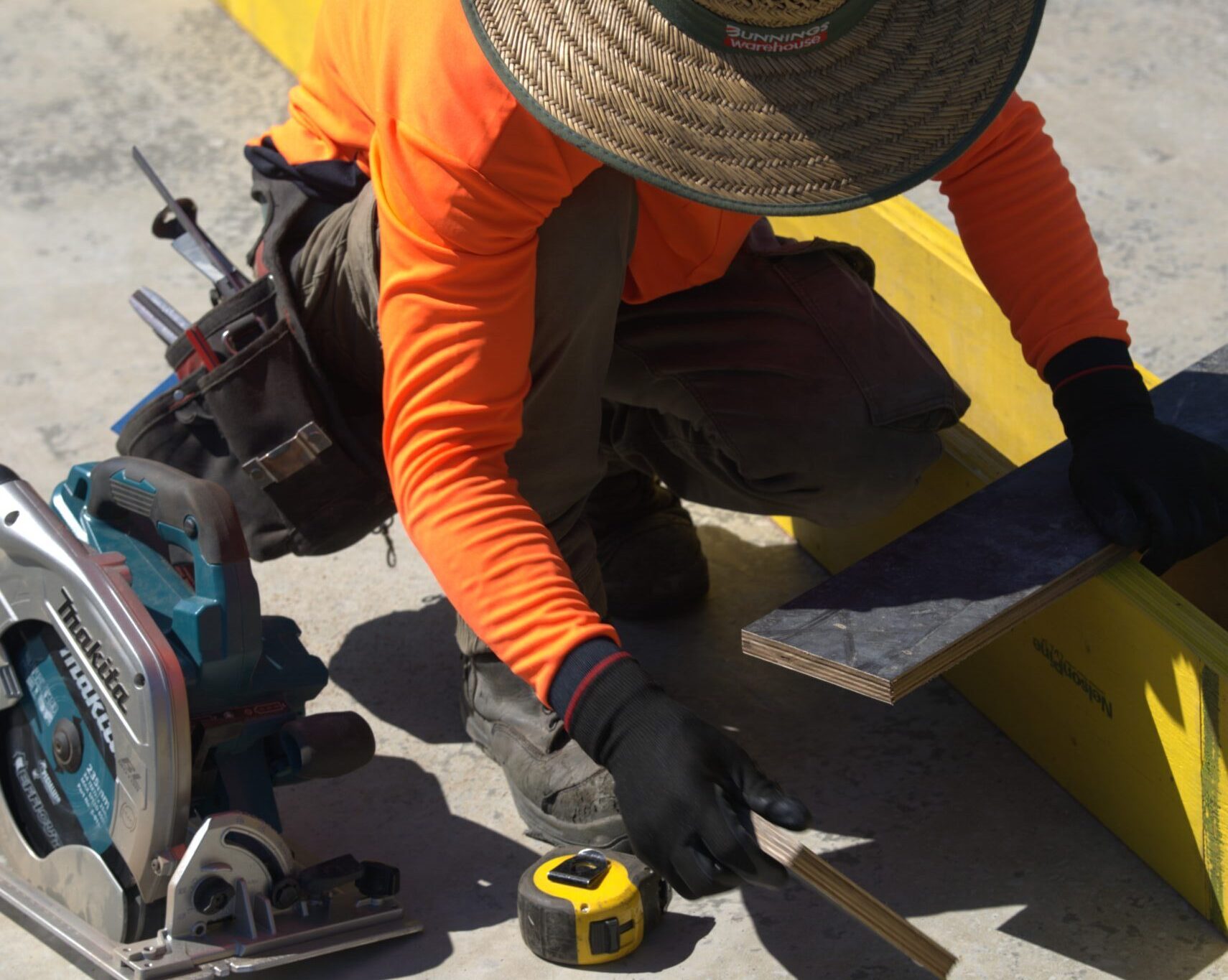 A person wearing a straw hat and orange long-sleeve shirt measures a wooden plank on a concrete surface, surrounded by a tape measure, power saw, and blue hose.