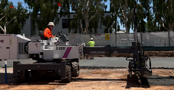 A construction leader servicing Southeast Queensland and New South Wales operates a white machine labeled "S-15R" on a dirt site, smoothing freshly poured concrete as another worker in a yellow vest stands nearby. Trees and a building are visible in the background.