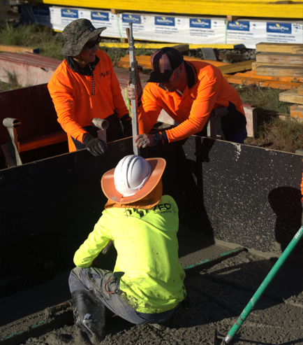 Three experienced construction industry leaders in brightly colored shirts and helmets are working with wet concrete at a building site. Two are standing and holding tools, while one is kneeling inside a formwork structure.